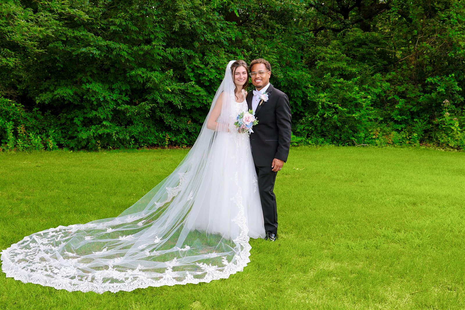 Bride and groom on a green lawn with the bride’s lace veil spread beside them